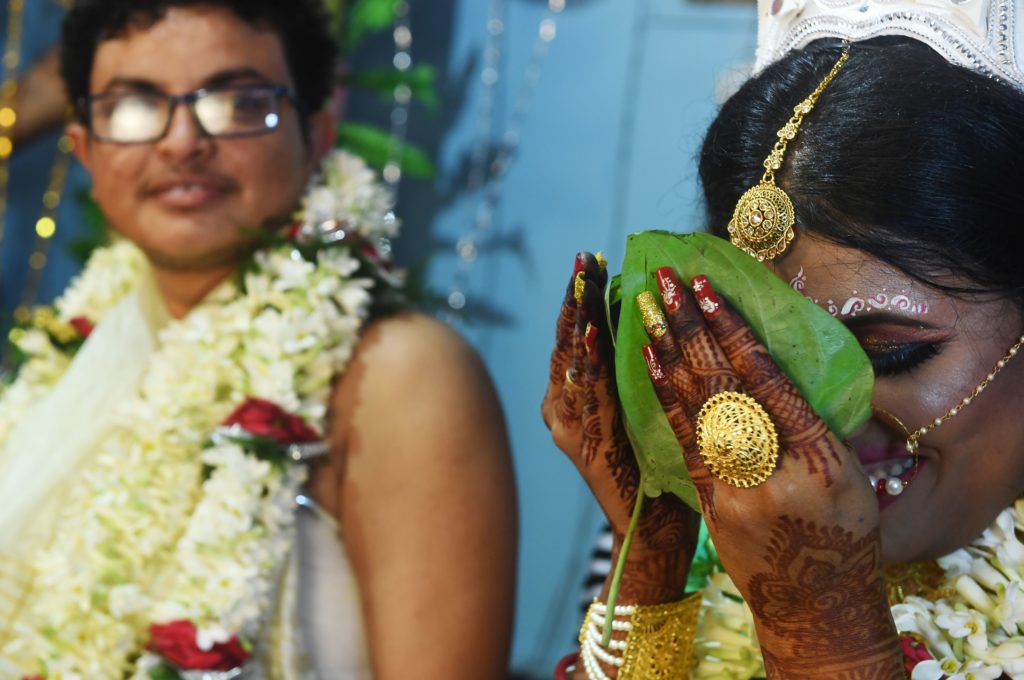 A trans bridge and groom performing marriage rituals, she is holding a leaf to her face as he looks on smiling