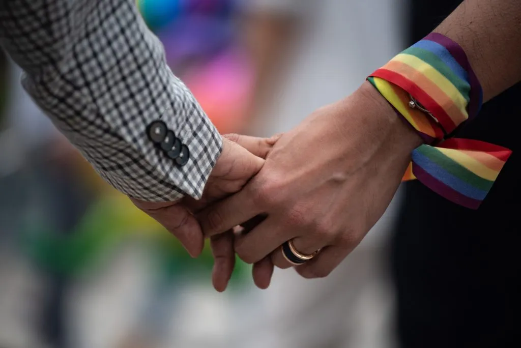 A close-up of a same-sex couple holding hands, one has a rainbow ribbon wrapped around their wrist