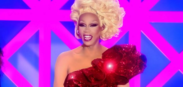 RuPaul in a one-shouldered sequinned red dress and a blonde updo, smiling behind the Drag Race UK judging table