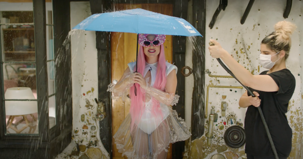 Lady Gaga holding an umbrella as her assistant pours water from a hose on her, or a work of art? (Screen capture via YouTube)