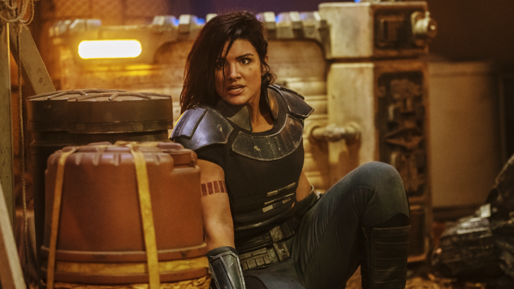 Gina Carano as Cara Dune in The Mandalorian in a body-armour style outfit, hiding behind a barrell