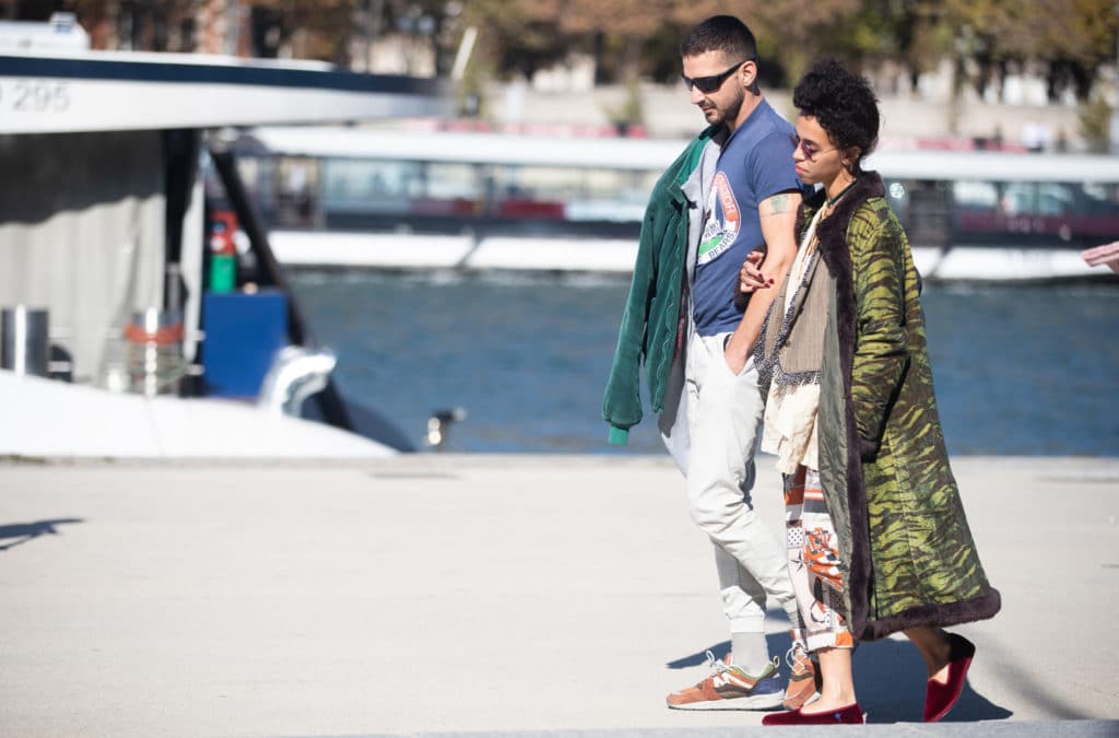 Shia LaBeouf and FKA Twigs walk along a pier with a boat behind them