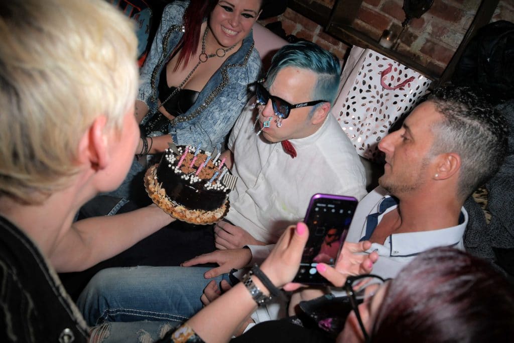 Michael Alig with blue hair and wearing sunglasses blows out the birthday candles of a cake