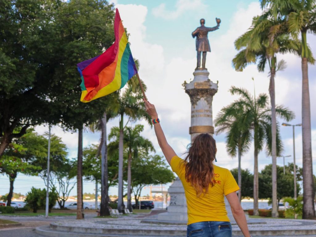 Linda Brasil waves an LGBT+ Pride flag with her back towards the camera in a yellow t-shirt