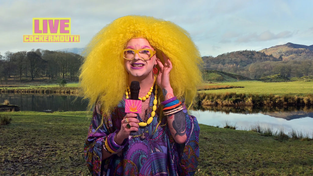 Ginny in a curly yellow wig and a paisley kaftan presenting the weather