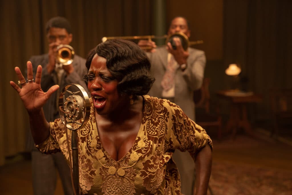 Ma Rainey (Viola Davis) singing into an old-fashion mic, with Levee (Chadwick Boseman) and Cutler (Colman Domingo) playing brass behind her