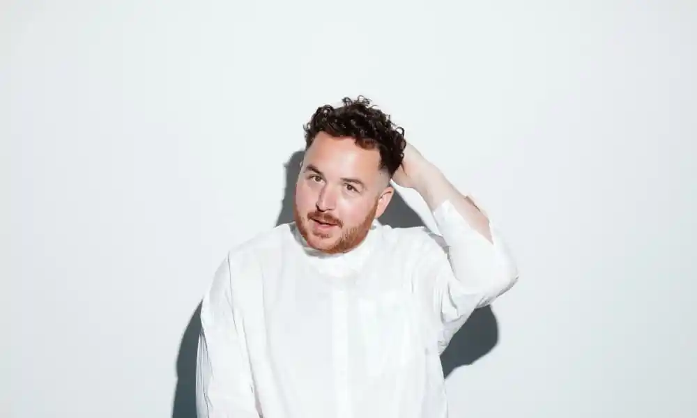Scottee with his hand behind his head in a white shirt against a white background