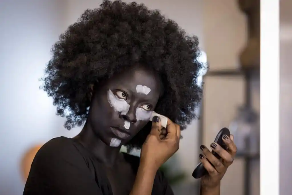 Glow Up's Dolli, a Black woman, who has painted her face a darker shade of brown, and is applying white stripes