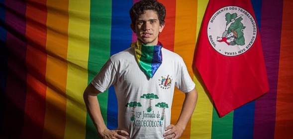 Lindolfo Kosmaski stands with his hands on his hips against the LGBT+ Pride and Landless Workers' Movement flags