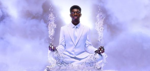 Lil Nas X sits in a white suit on a pathway made of ice floating in the skies