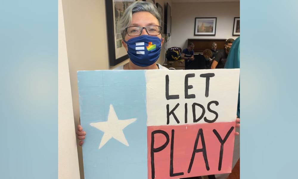 Picture of Hillary Moore-Embry who is a parent fighting anti-trans legislation in Texas, they hold up a sign that reads 'let kids play'