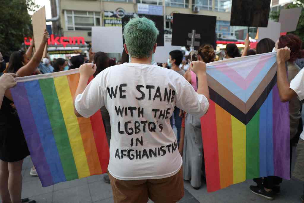 Person stood with their back to the camera. The back of their top reads: "We stand with LGBTQs in Afghanistan". They're holding a rainbow Pride flag and the Progress Pride flag