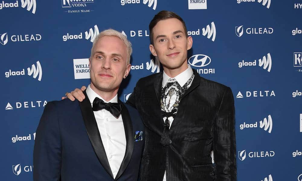 Adam Rippon and Jussi-Pekka Kajaala are dressed in dark suits and lighter shirts as they smile at the camera while standing in front of a blue background with various brand logos on it
