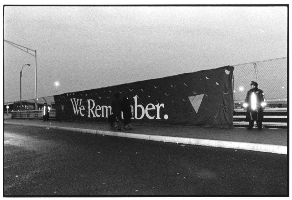 A huge "We Remember" banner handing on a fence flanked by police officers during a demonstration against the killing of gay man in February 1990 on Staten Island, New York