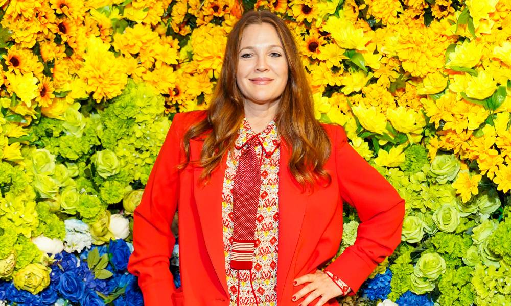 Drew Barrymore wears a patterned top and a red jacket as she stands in front of a rainbow-coloured background made up of flowers