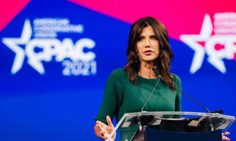 Kristi Noem wears a green outfit and stands at a podium in front of a red and blue background