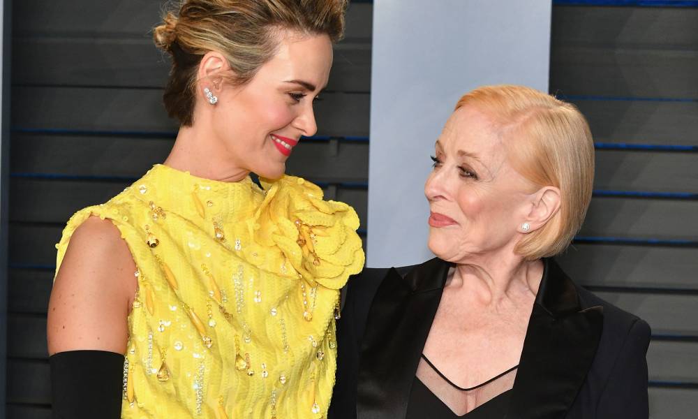 Sarah Paulson and Holland Taylor smile at each other. Paulson is dressed in a yellow dress with long black gloves while Taylor wears a black top and black jacket
