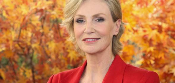 Jane Lynch, a white woman with blonde hair, wears a black shirt and red jacket while standing in front of orange leaves