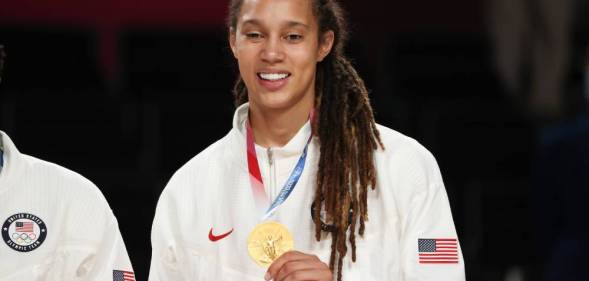 Brittney Griner, a basketball player, holds up her Olympic gold medal while wearing a white Team USA jacket