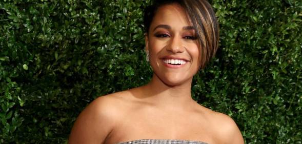 Ariana DeBose smiles at the camera while standing in front of a background filled with greenery