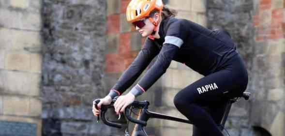 Trans cyclist Emily Bridges rides a black bicycle in front of a brick building