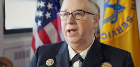 Dr Rachel Levine speaks to a person off camera while wearing a blue uniform with an American flag in the background