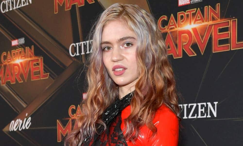 Grimes stares at the camera while wearing a black and red outfit