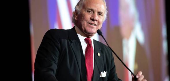 South Carolina governor Henry McMaster wears a white shirt, red tie and dark suit jacket as he speaks into a microphone on stage