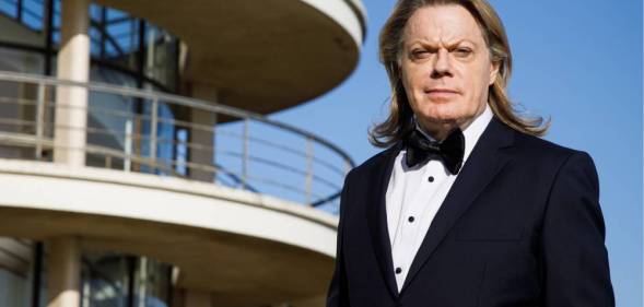 Eddie Izzard is see wearing a black suit jacket, white shirt and black bow tie