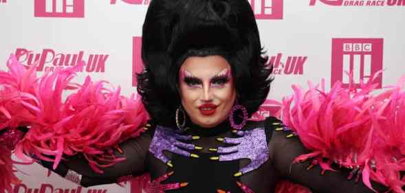 Drag Race UK star Choriza May stares at the camera while wearing a black wig, black outfit with pink and purple hands on it