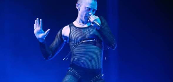 Years & Years singer Olly Alexander sings into a microphone while wearing a black mesh bodysuit and being lit by a blue light