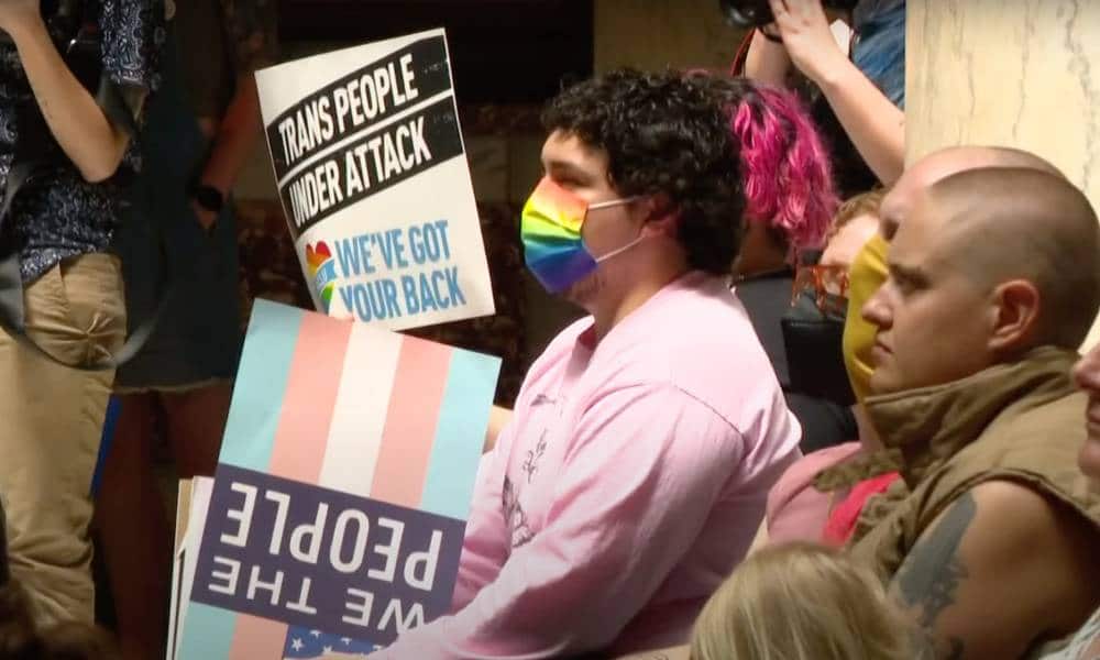 A person wearing a rainbow face mask holds a sign that in the colour of the trans flag while a sign reading 'trans people are under attack' is seen in the background