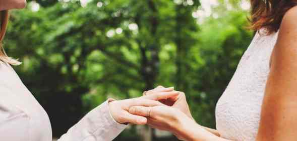 Two female presenting people, who are presumed to be in a lesbian or queer relationship, face each other while wearing wedding dresses as one person puts a ring on the other's hand