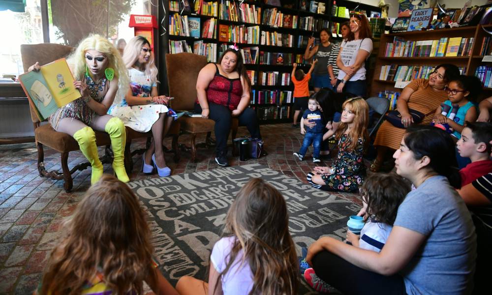 Two drag artists sit in front of a crowd of children and adults. One performer is reading from a book while another performer is smiling at the crowd