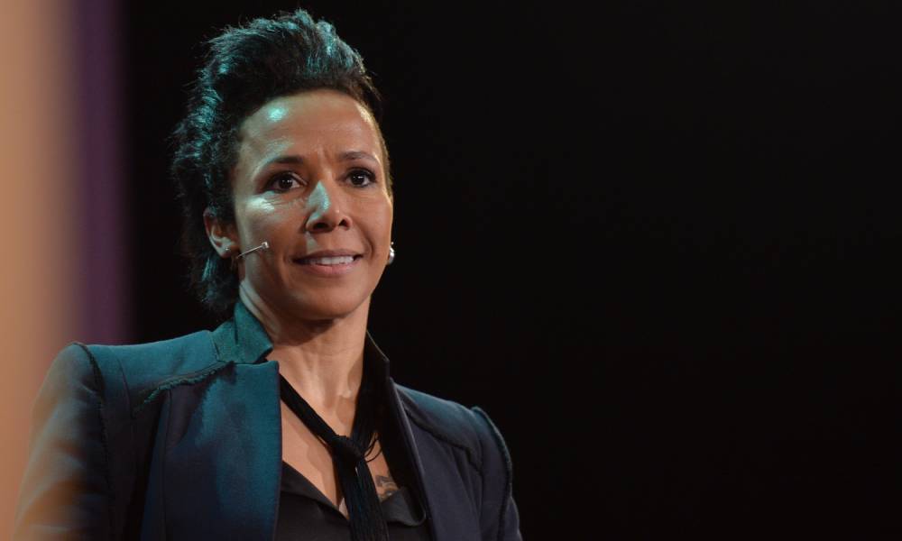 Dame Kelly Holmes wears a black top, black blazer and matching tie as she speaks to people off camera