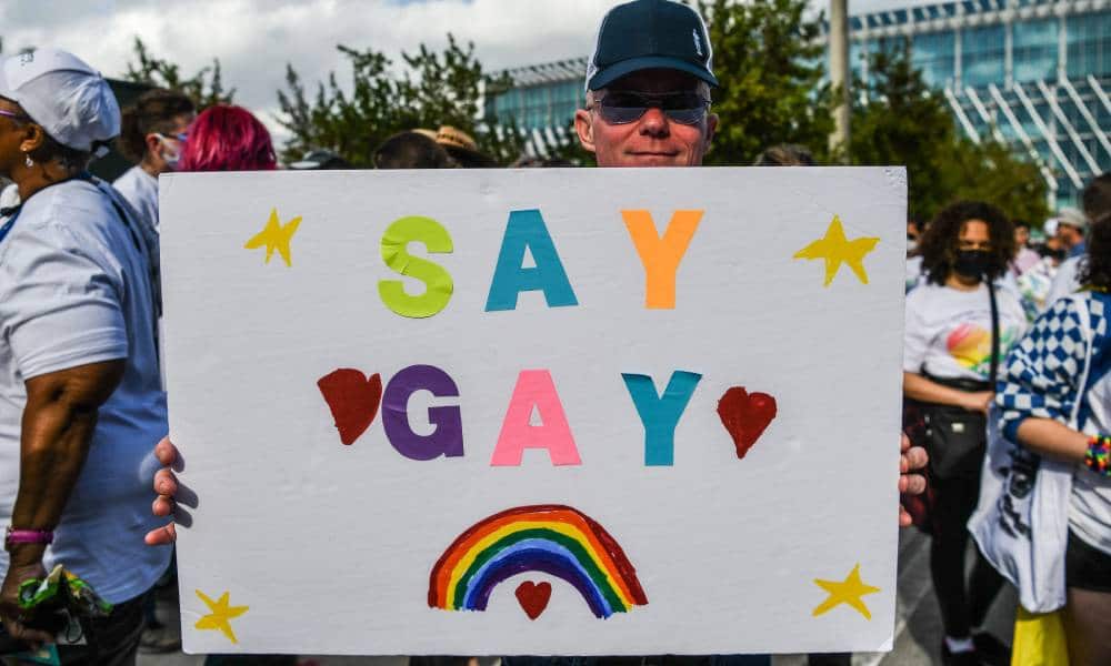 A person wearing a baseball cap and sunglasses holds up a sign that reads 'Say gay' in colourful letters and a tiny rainbow underneath
