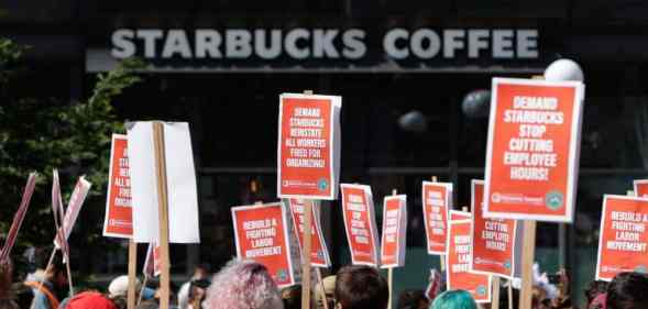 People in a crowd hold up various orange signs with white writing in support of Starbucks unionising with a white Starbucks logo from a store is seen in the background