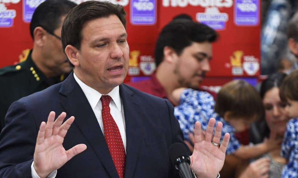 Florida governor Ron DeSantis wears a white button up shirt, red tie and dark suit jacket as he stands at a podium and speaks into a microphone. He is gesturing with both his hands up in the air