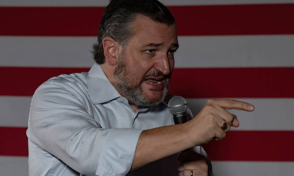 Texas senator Ted Cruz wears a white button up shirt as he speaks into a microphone and points with a finger on his other hand at something off screen with the red and white stripes of the American flag seen in the background