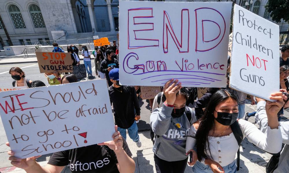 Students participate in a school walk-out and protest in front of City Hall to condemn gun violence. One person holds up a sign reading 'We should not be afraid to go to school!' Another person has a sign reading 'End gun violence' while a third person, who is wearing a face mask, holds up a sign that says 'Protect children not guns'