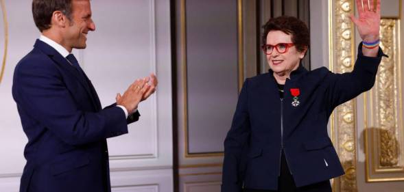 French president Emmanuel Macron wears a dark suit and white button up shirt as he stands sideways and claps for Billie Jean King, who is dressed in a black outfit with a navy blue jacket on top. She has the France's Legion of Honour award pinned to her chest and raises her left arm in the air, and she is wearing a rainbow bracelet around her wrist