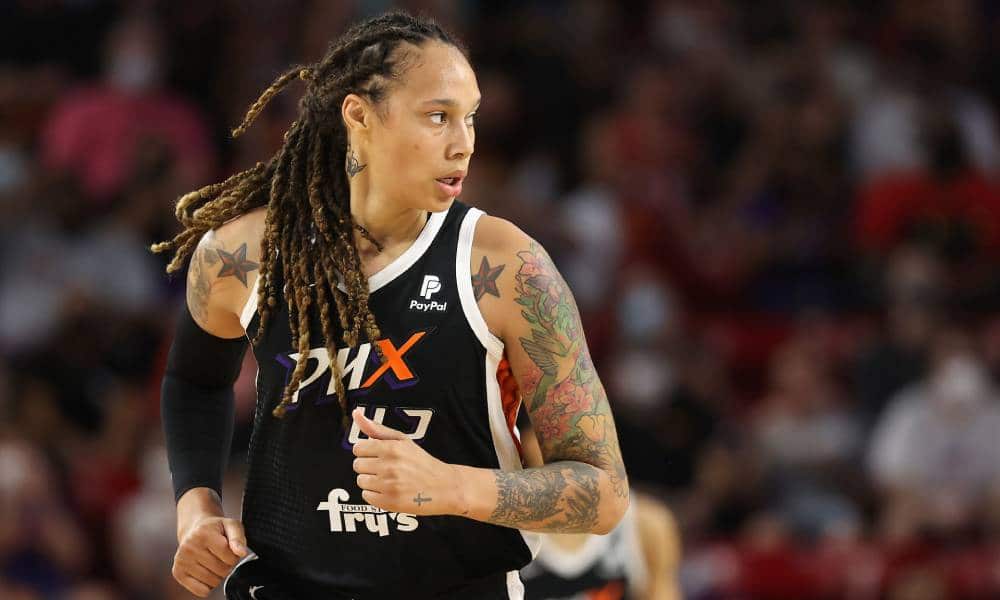 Brittney Griner wears a black Phoenix Mercury jersey with white panelling as she runs down a basketball court. Her hair is styled in locks and her arm has tattoos on it and she looks to the side at someone else