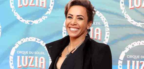 Dame Kelly Holmes wears a black top and black jacket as she smiles at the camera and stands in front of a blue background