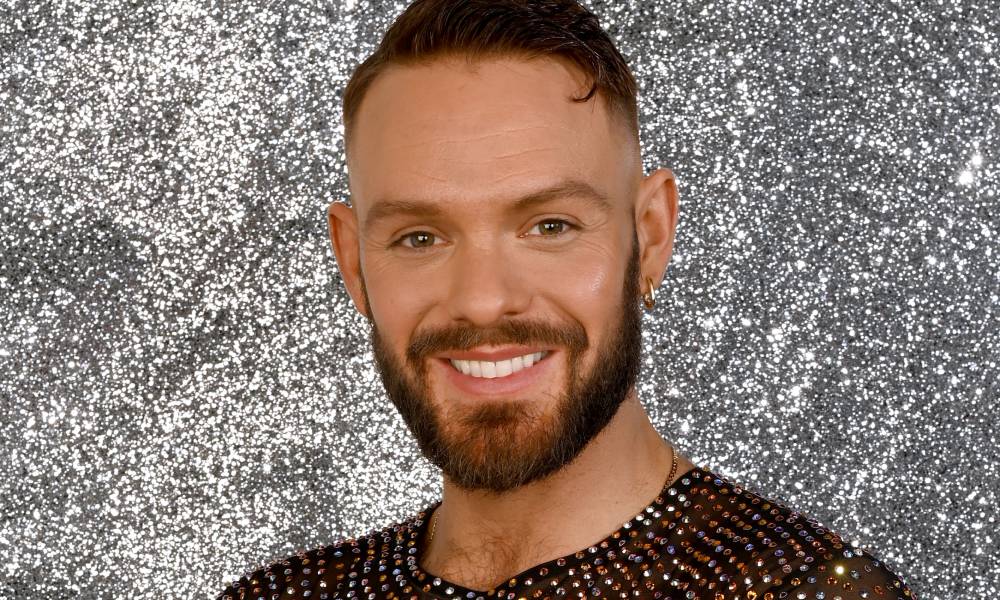 John Whaite wears a sparkly top as he smiles at the camera and stands in front of a silver sparkly background