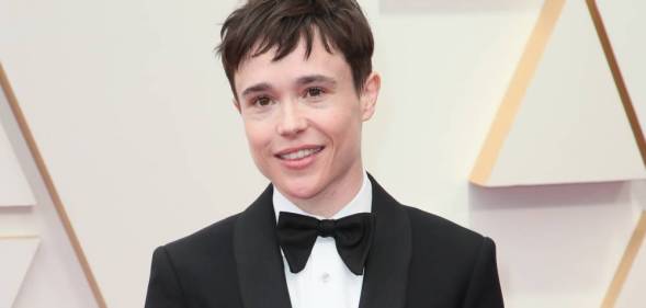 Elliot Page smiles for the camera while wearing a bow tie, white button up shirt and black suit jacket and standing in front of a pale off-white background