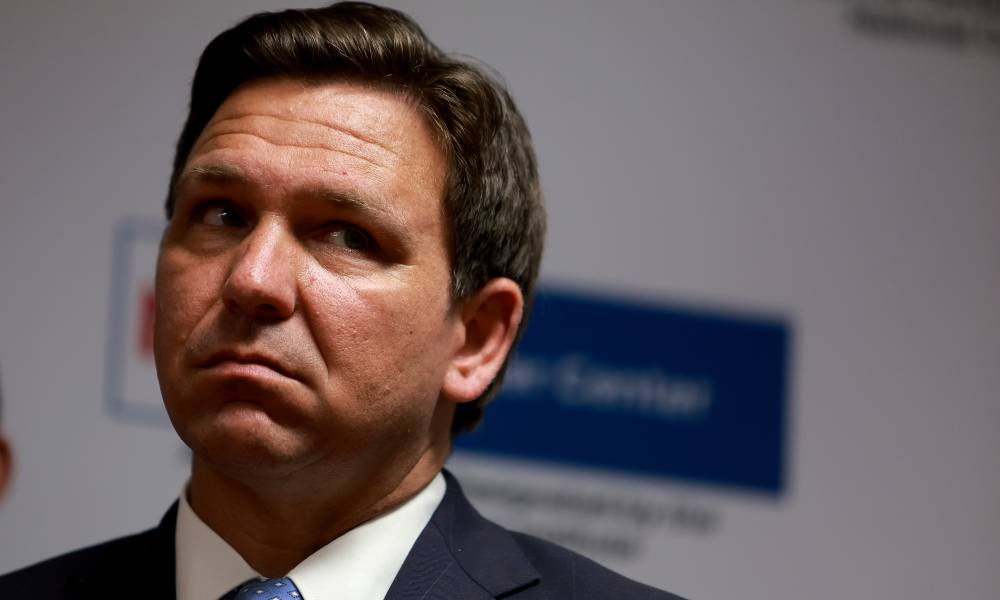 Florida governor Ron DeSantis wears a white button up shirt, light blue tie and dark blue jacket as he stares at someone off camera