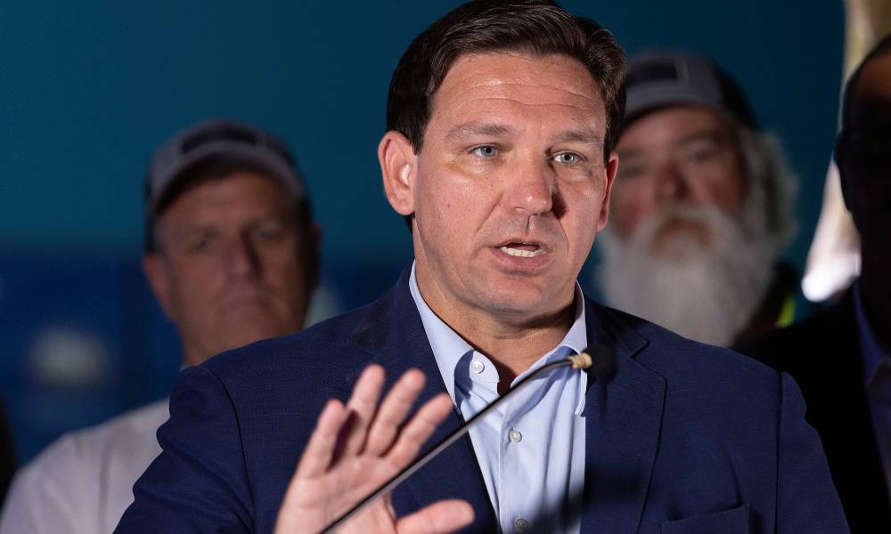 Florida governor Ron DeSantis wears a light blue button up shirt and dark blue suit jacket as he speaks into a microphone in front of a crowd of people. He is gesturing with one hand as several people stand behind him