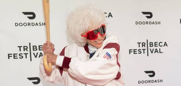 Maybelle Blair, an original All-American Girls Professional Baseball League player, wears a white and red coat with dark red rimmed glasses and she poses for the cameras with a baseball bat as if she is ready to hit a ball