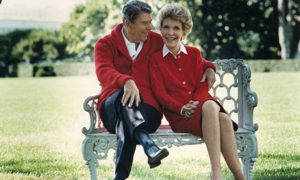Former president Ronald Reagan sits on a bench outside while wearing a red coat, white shirt and black trousers. He has his arm around his wife, Nancy Reagan, who is wearing a matching outfit. Ronald is staring at her while Nancy is looking at the camera.