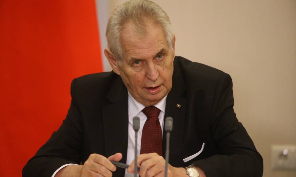 Czech president Miloš Zeman wears a white button up shirt, red tie and black jacket as he speaks into two grey microphones while tilting his head down